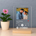 A clear acrylic music player plaque on a light wood base, displaying a custom Spotify-style player interface with the song 'Tere Hawaale' by Anjit Singh and a photo of a couple. The plaque is styled on a light beige surface next to a potted pink flower against a textured gray wall.