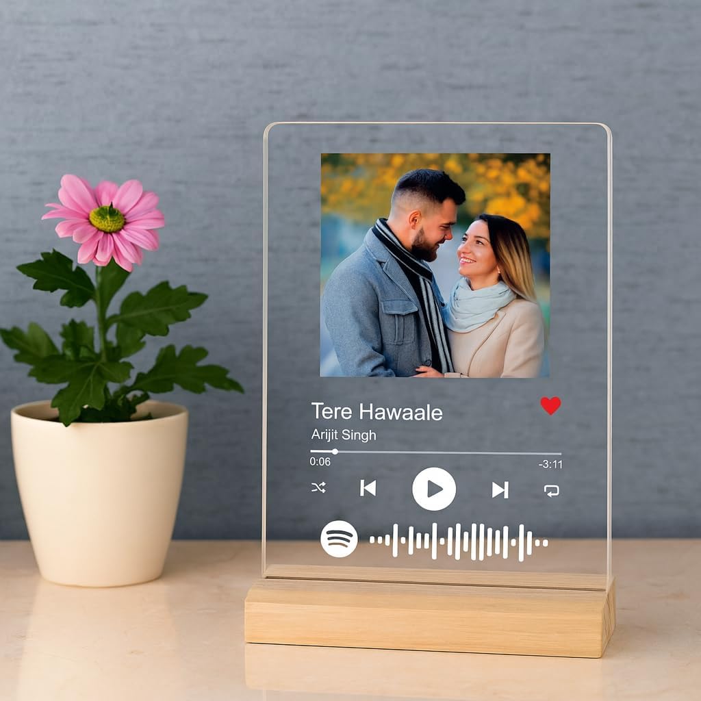 A clear acrylic music player plaque on a light wood base, displaying a custom Spotify-style player interface with the song 'Tere Hawaale' by Anjit Singh and a photo of a couple. The plaque is styled on a light beige surface next to a potted pink flower against a textured gray wall.